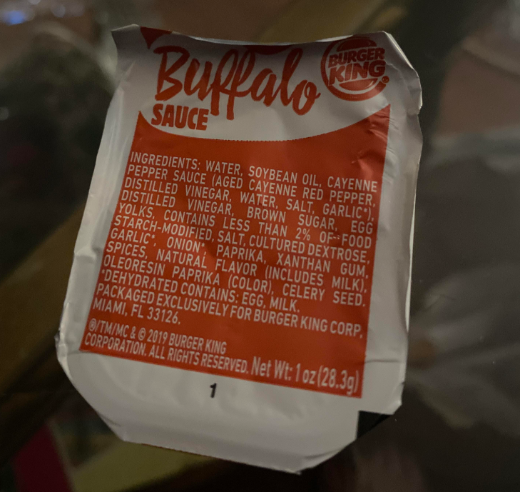 A close-up of a Burger King Buffalo Sauce packet lid, featuring the ingredients list and product information for this popular fast-food sauce in black and orange text on a white and orange background.