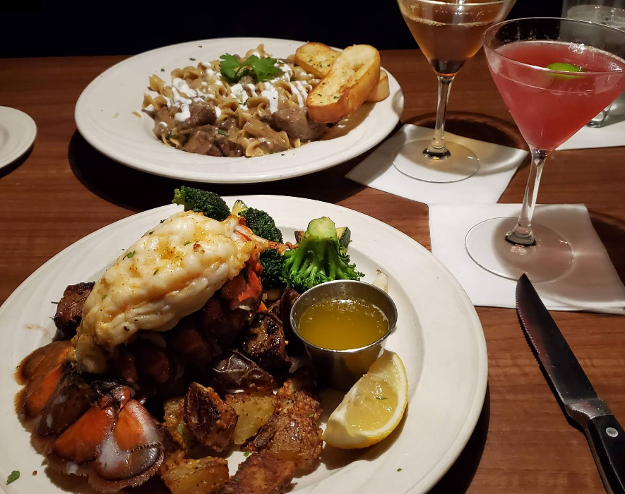 A plate with lobster tail, roasted potatoes, broccoli, lemon wedge, and melted butter sits in front of a pasta dish with beef and toast at a steakhouse on the West Coast, alongside two cocktails on a wooden table in a restaurant setting.