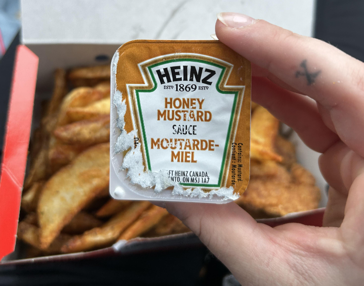 A hand holds a frost-covered Heinz honey mustard sauce packet, one of the classic fast-food sauces, above a box of crispy potato wedges.