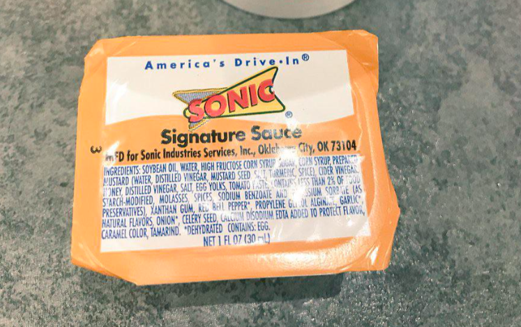 A packet of Sonic Signature Sauce, one of the popular fast-food sauces, sits on a gray surface. The label displays the Sonic logo, ingredient list, and serving size of 1 fl oz (30 ml).
