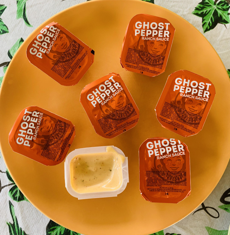 Six Ghost Pepper Ranch fast-food sauce packets arranged on a yellow plate, with one packet open displaying creamy sauce inside. The plate rests on a table covered by a green and white patterned tablecloth.