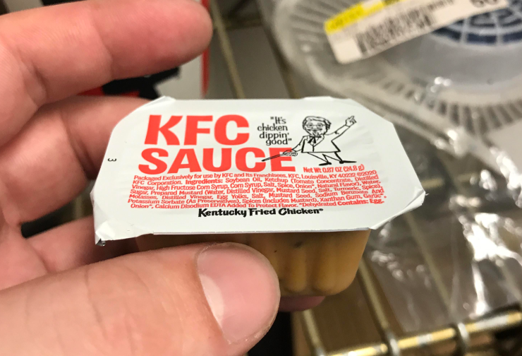 A hand holds a small plastic container of KFC Sauce, one of the iconic fast-food sauces with a red and white label featuring Colonel Sanders and the phrase "It's chicken dippin' good." Blurred shelves and other items fill the background.