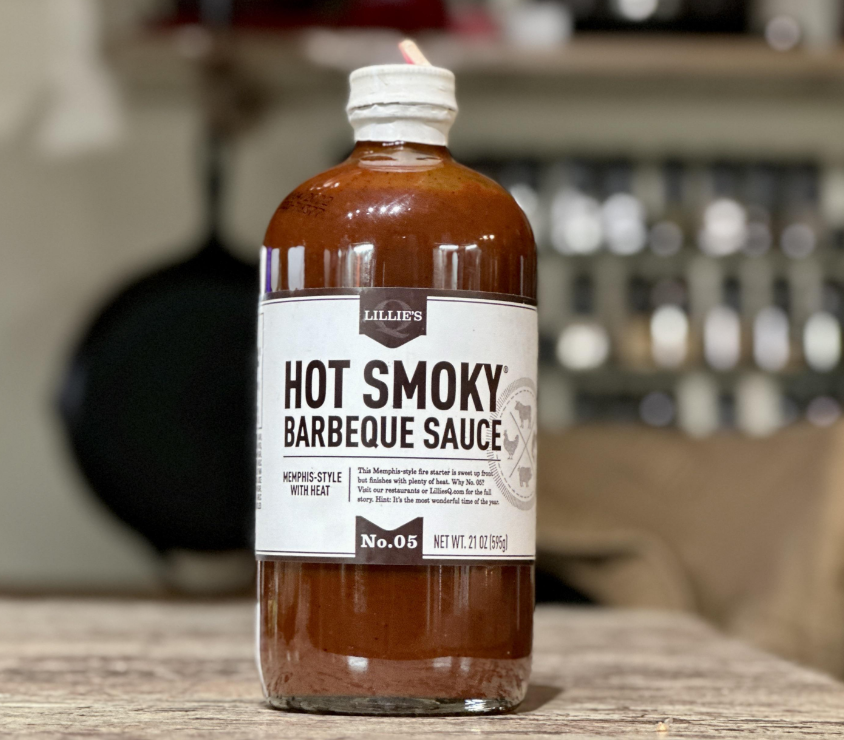 A glass bottle of Lillie’s Hot Smoky Barbeque Sauce, inspired by Memphis-style fast-food sauces with heat, sits on a wooden surface. The label highlights its spicy kick and 21 oz (595g) size against a blurred background.