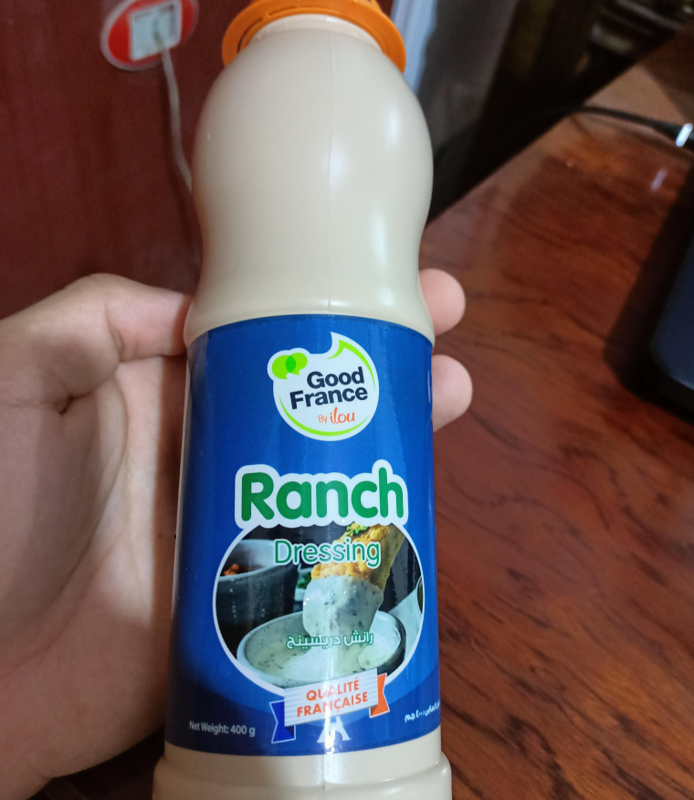 A hand is holding a bottle of "Good France" ranch dressing—a popular choice among fast-food sauces. The blue label, with salad imagery and English-Arabic text, complements the vibrant orange bottle cap.