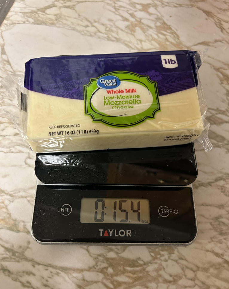 A block of Great Value whole milk low-moisture mozzarella cheese sits on a digital kitchen scale, displaying a weight of 0.154 kilograms. The cheese package is labeled as 1 lb (16 oz, 453g).