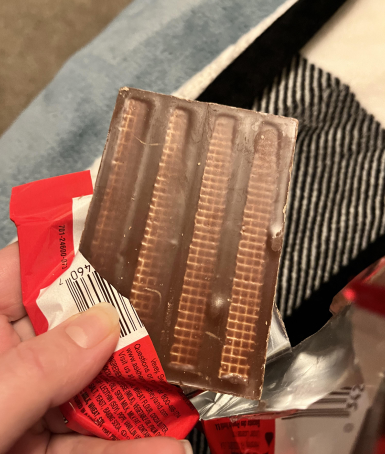 A hand holds a partially unwrapped chocolate bar, revealing wafer layers visible through the chocolate. The wrapper is red and silver, and a barcode is shown on the packaging. A striped fabric is in the background.