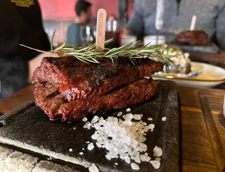 A close-up of grilled steak slices stacked on a stone plate, topped with rosemary and a "medium" label. Coarse sea salt is scattered nearby, evoking the signature style of a steakhouse on the west coast. Blurred food and people fill the background.