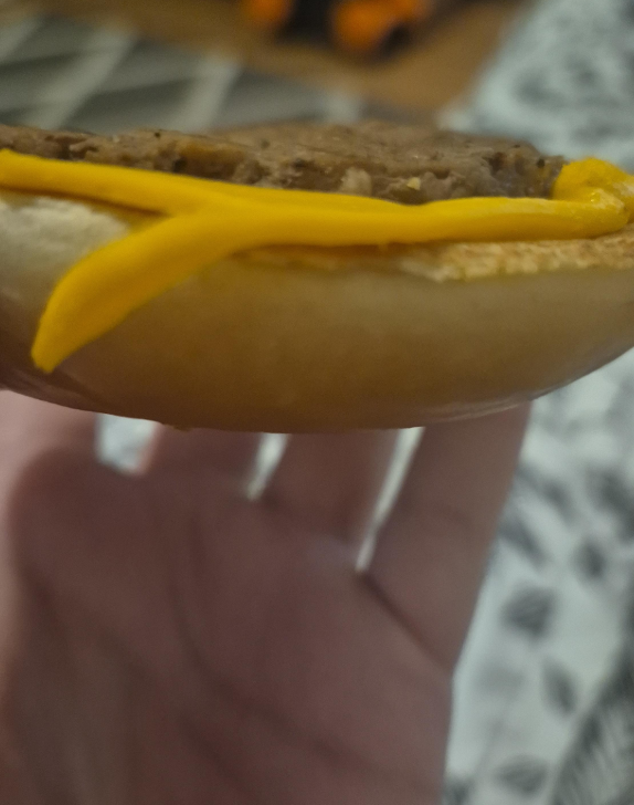 A close-up side view of a hand holding a hamburger bun with a slice of cheddar cheese and beef patty inside, with the sandwich partially open and background out of focus.