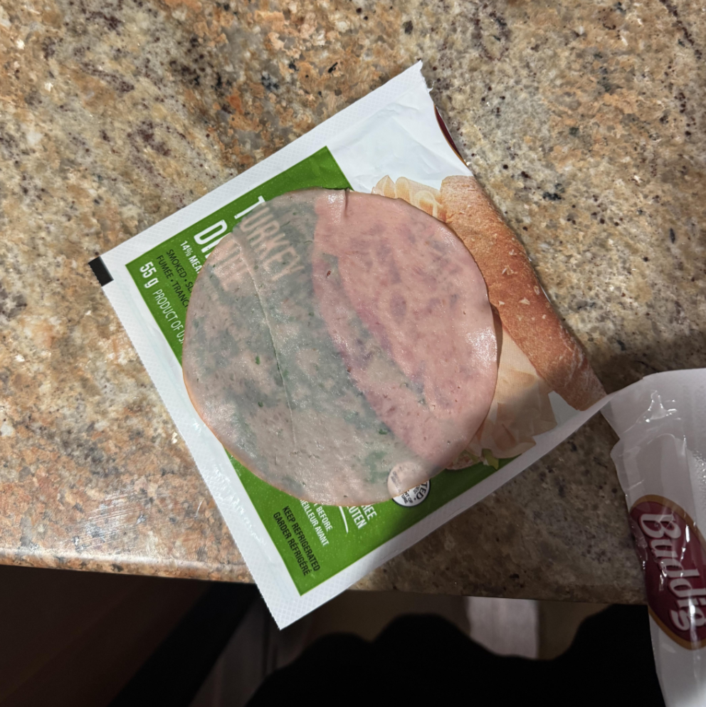 A slice of turkey lunch meat with greenish mold sits on partially opened packaging on a speckled kitchen countertop. A bread bag with the word "Bailey's" is visible at the edge of the image.