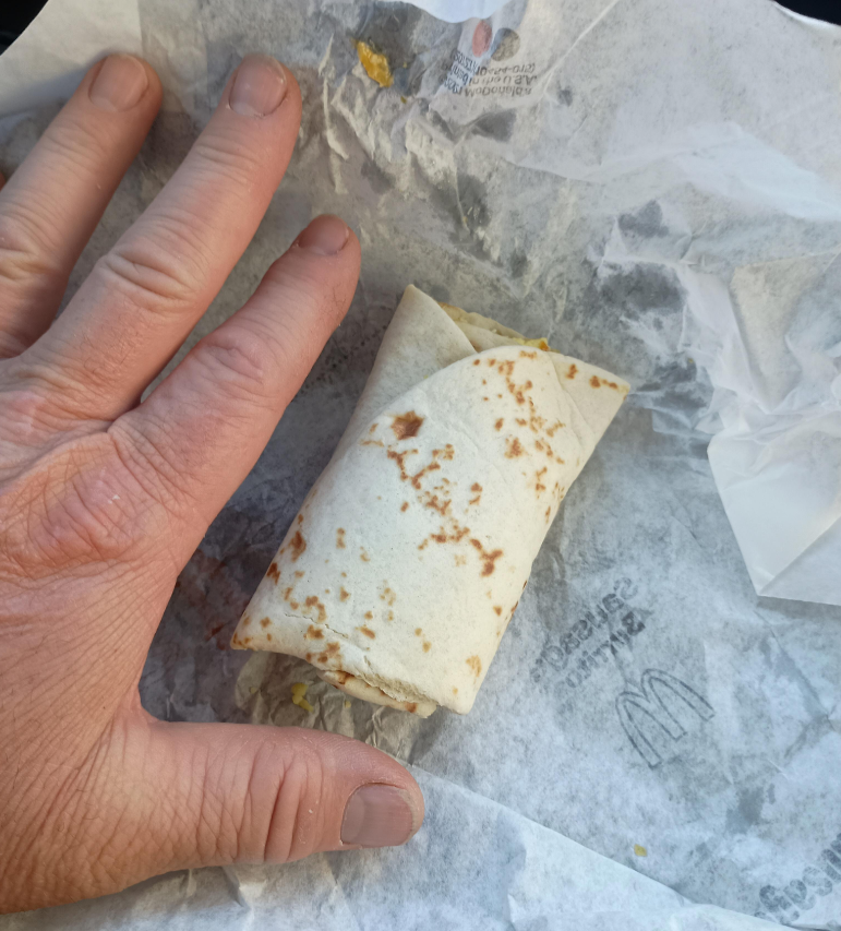 A hand next to a small folded tortilla wrap placed on McDonald's branded sandwich paper, highlighting the wrap’s compact size.