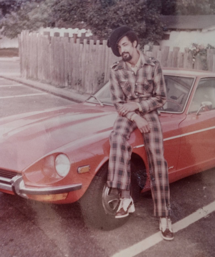 A man in a plaid suit and beret sits on the hood of a classic red car in a parking lot, smiling at the camera. A wooden fence and trees are in the background. The photo has a vintage feel.