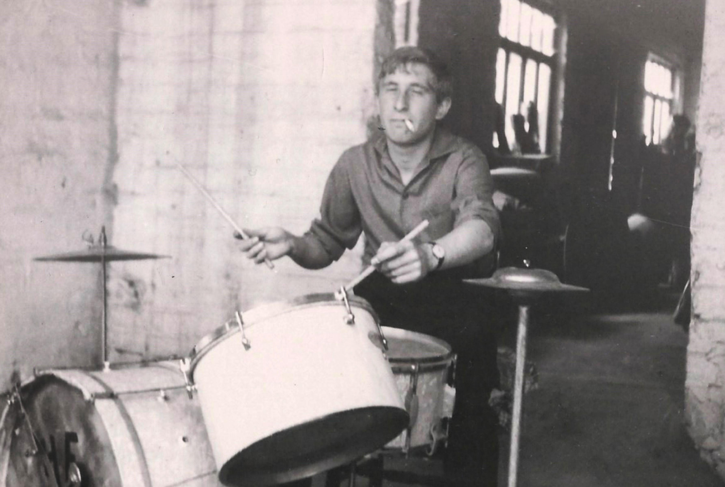 A young man with a cigarette in his mouth plays a drum set in a narrow, dimly lit room with brick walls and large windows. The image is black and white, giving it a vintage feel.