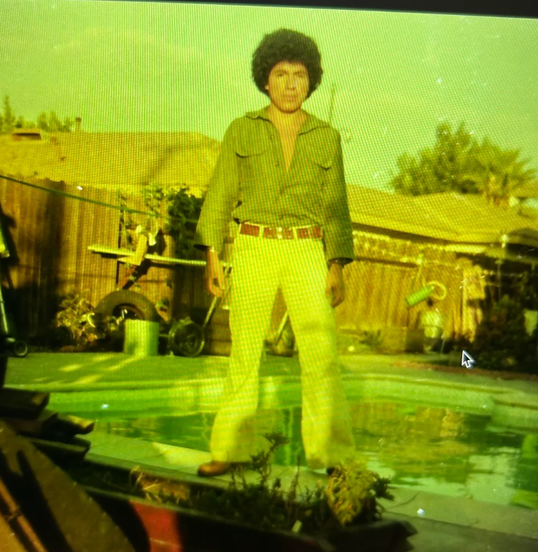 A person with curly hair and a green shirt stands by a backyard swimming pool, surrounded by plants and a wooden fence, with houses and trees in the background. The image has a yellow-green tint.