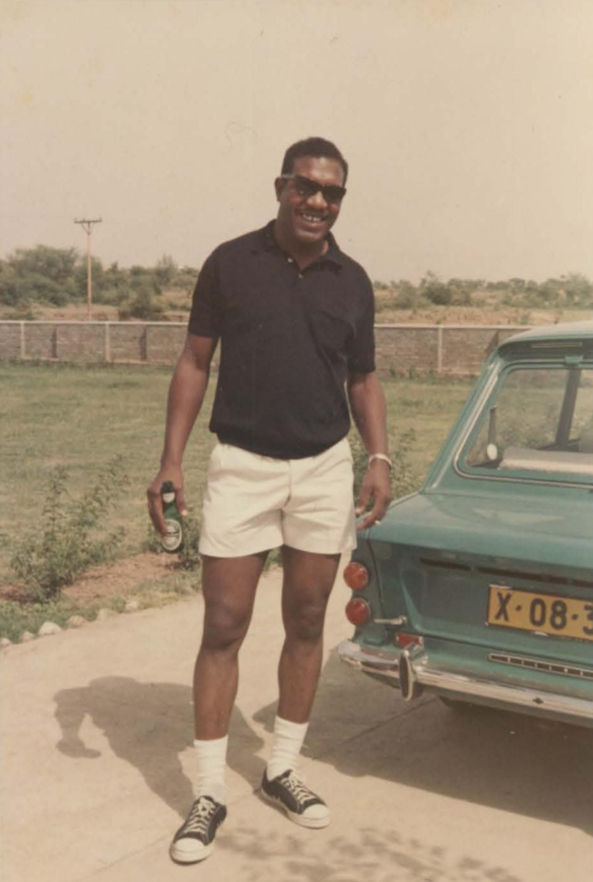 A man wearing sunglasses, a navy polo shirt, white shorts, and Converse sneakers stands smiling beside a vintage green car, holding a drink, with grass and a fence visible in the background.