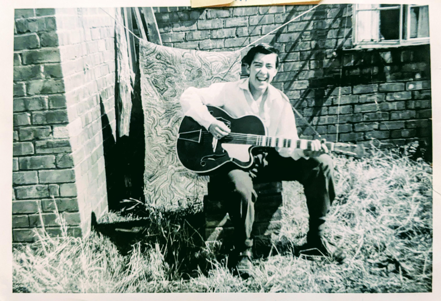 A man sitting on bricks near a brick wall, playing an acoustic guitar and smiling. There is a patterned blanket hanging behind him and tall grass in the foreground. The photo is in black and white.