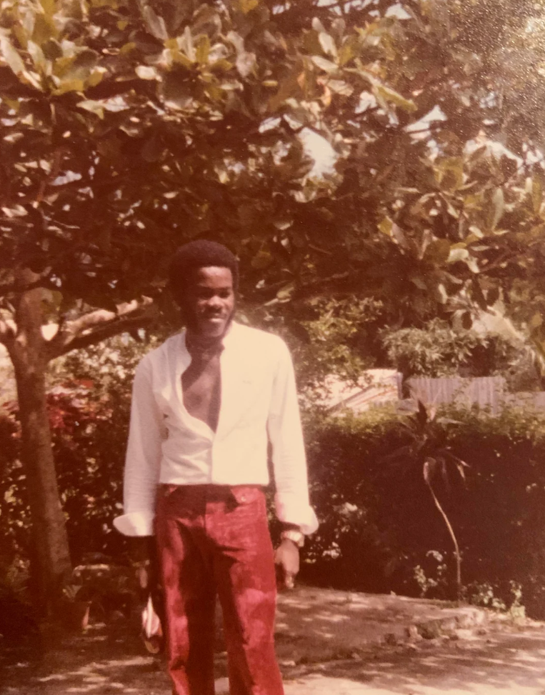 A man stands outdoors under a leafy tree, wearing a white shirt partially unbuttoned and red pants. Sunlight filters through the branches, and lush greenery surrounds him. He appears to be smiling.