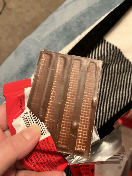 A hand holds an unwrapped chocolate bar with four visible wafer segments, partially removed from its red and silver packaging. A striped fabric is in the background.