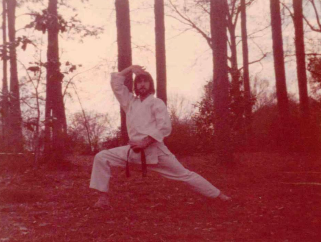 A person in a karate uniform and black belt performs a martial arts pose outdoors in a wooded area, surrounded by tall trees and scattered leaves on the ground.