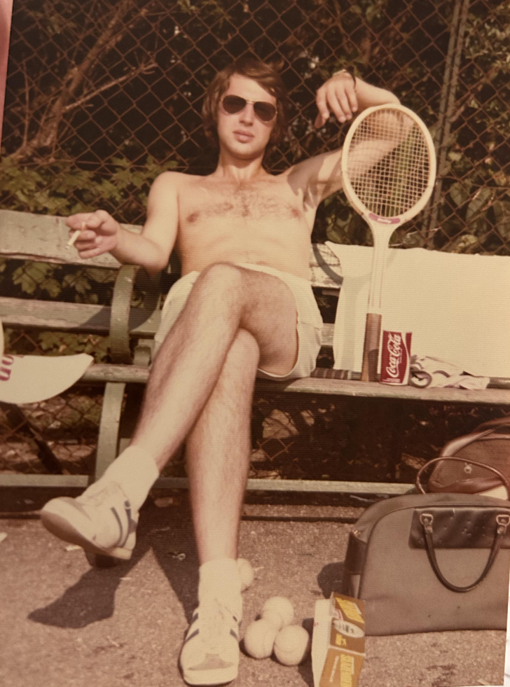 A young man wearing sunglasses and shorts relaxes shirtless on a park bench, holding a cigarette. A tennis racket, tennis balls, a can of Coca-Cola, and a bag are nearby. A chain-link fence and greenery are in the background.