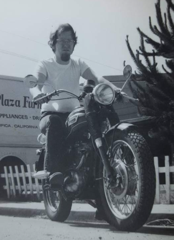 A man wearing glasses and a white t-shirt sits on a vintage motorcycle near a white picket fence. A moving truck labeled "Plaza Furniture" is in the background. The image is in black and white.