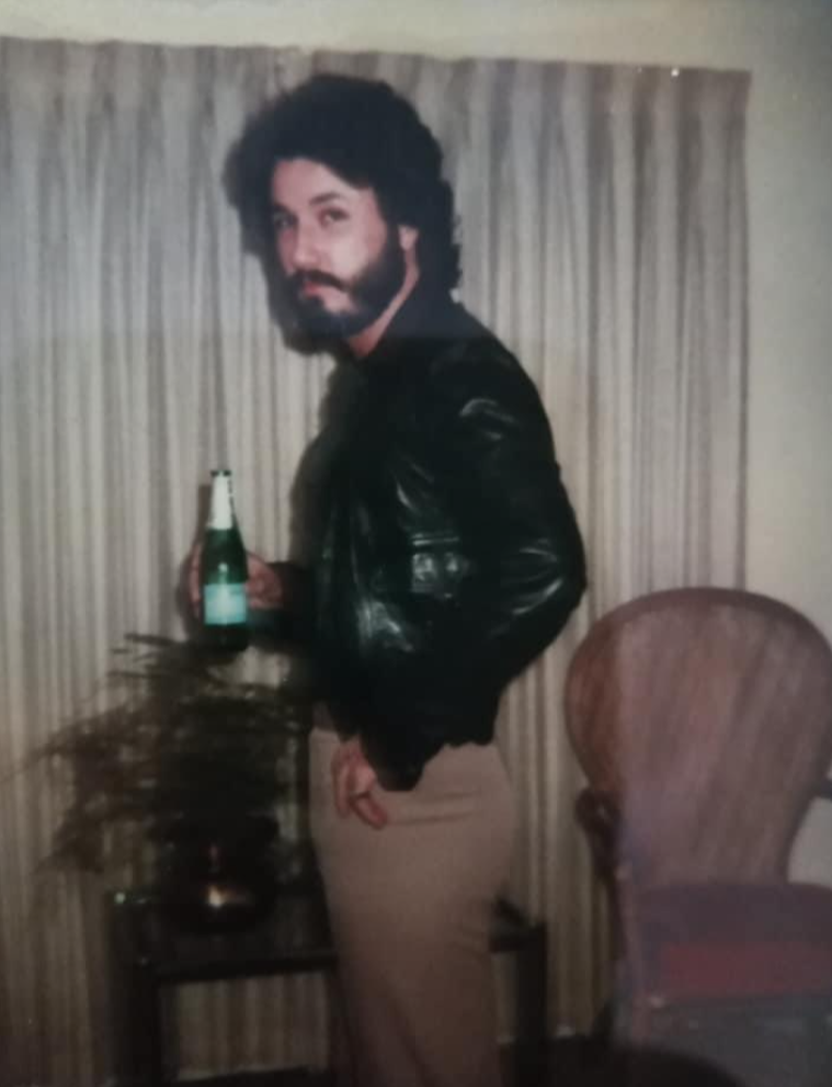 A man with dark, curly hair and a beard stands indoors, wearing a black leather jacket and tan pants. He holds a green bottle and poses beside a small table with a plant, with beige curtains and a chair in the background.