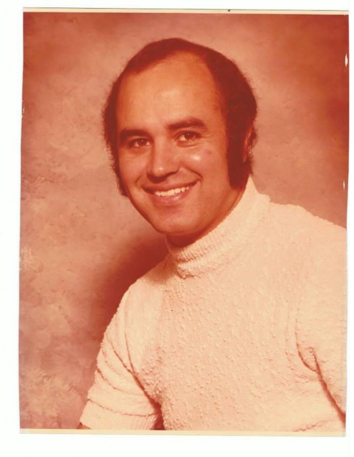 A man with dark hair and sideburns smiles at the camera, wearing a textured white turtleneck sweater, posed against a soft, brownish studio background. The photo has a warm, vintage tone.
