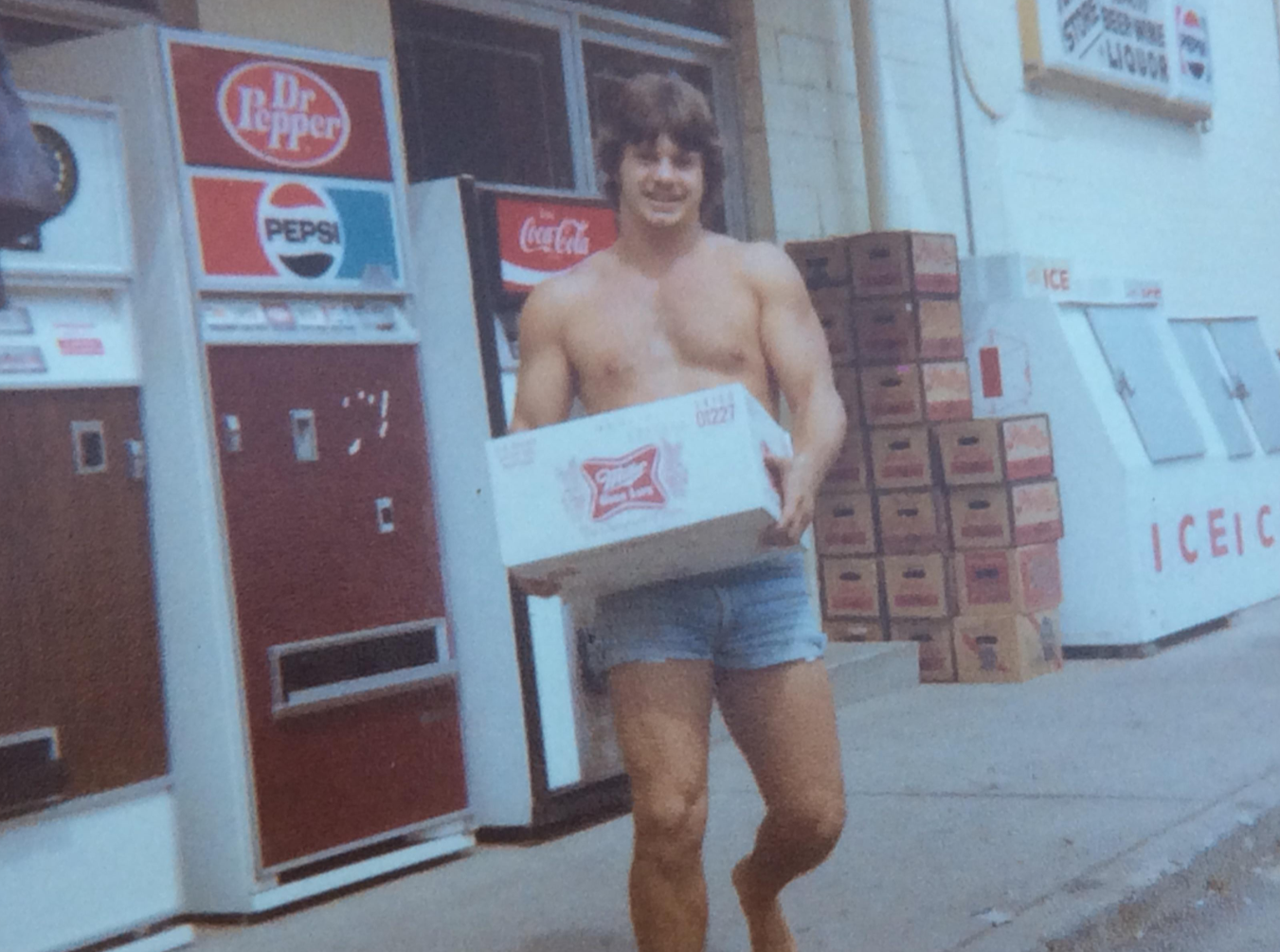A smiling, shirtless man in denim shorts carries a case of Budweiser outside a store with soda vending machines, ice freezers, and stacked boxes in the background.