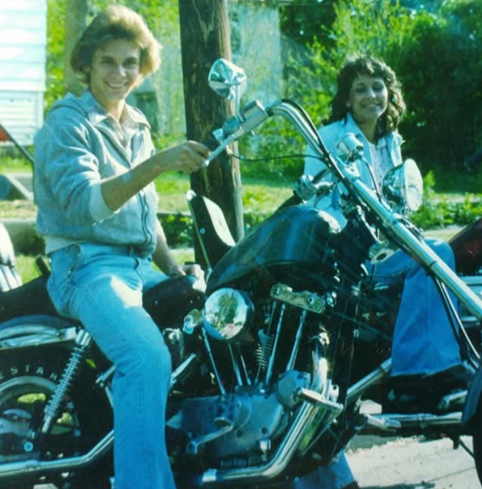 Two people sit on motorcycles outdoors, both smiling at the camera. The person in front wears a light gray hoodie and blue jeans, while the person behind wears a white jacket. Trees and greenery are visible in the background.