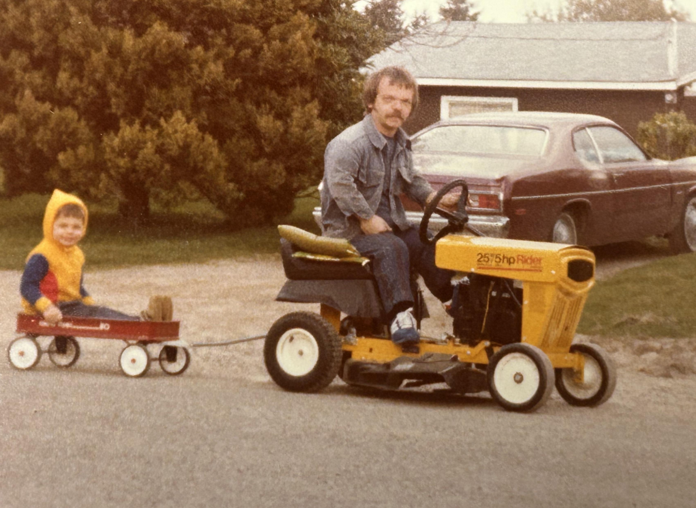 A man rides a yellow lawn tractor pulling a small red wagon with a smiling child in a yellow hoodie sitting inside. They are outside near trees and a parked car, in front of a house.