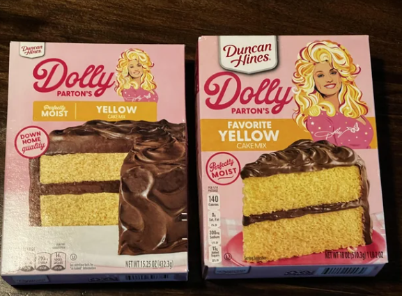 Two boxes of Dolly Parton's Duncan Hines yellow cake mix sit on a table. Both feature an illustration of Dolly Parton and an image of a yellow layer cake with chocolate frosting on the front.