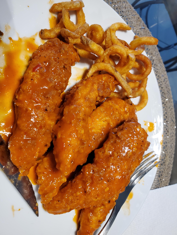 Four breaded chicken tenders covered in zesty orange wing sauces are served with a portion of curly fries on a white plate. A fork rests on the edge, ready for you to dig in.