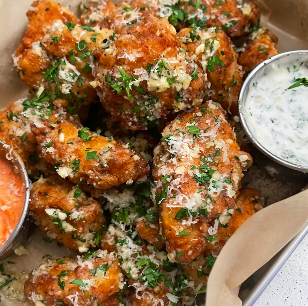 A close-up of crispy chicken wings garnished with grated cheese and chopped herbs, served with two flavorful wing sauces—one creamy white sauce with herbs and one bright orange sauce. The wings are arranged on parchment paper.