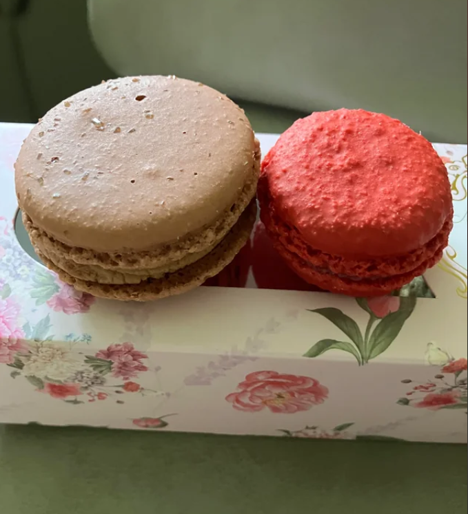 Two macarons, one light brown and one bright red, are placed side by side on a floral-decorated box. The background is softly blurred.
