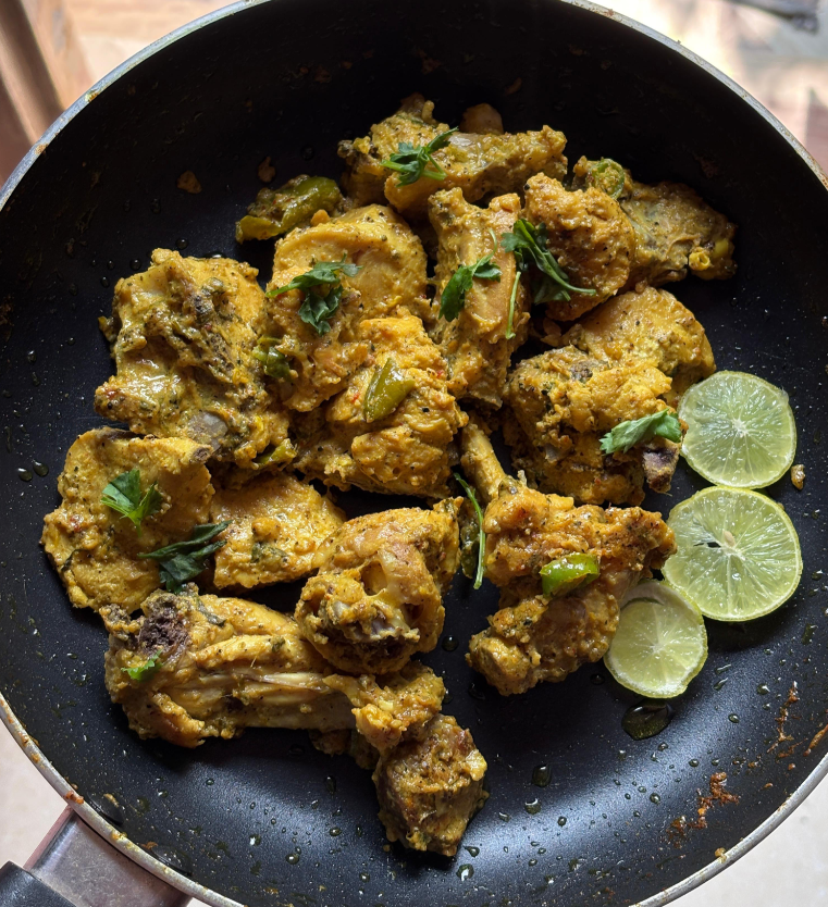 A pan of cooked chicken pieces marinated in spices and wing sauces, garnished with fresh coriander and accompanied by three slices of lime on the side.