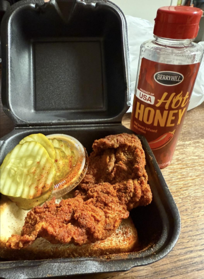 A black takeout container with fried chicken pieces on a slice of bread, a side of sliced pickles, and a condiment cup. A bottle of Berryhill hot honey wing sauce is placed next to the container on a wooden surface.