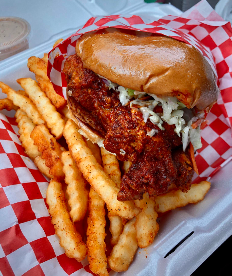 A takeout container with crinkle-cut fries and a spicy fried chicken sandwich topped with coleslaw on a toasted bun, served on red and white checkered paper—perfect for pairing with your favorite wing sauces.