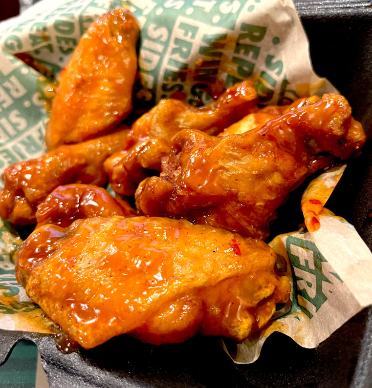 A close-up of crispy chicken wings coated in a glossy, spicy orange wing sauce, served in a black takeout container lined with branded paper.