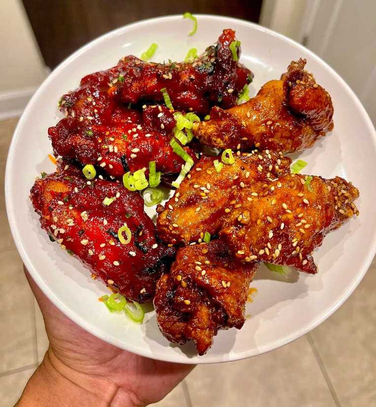 A hand holds a white plate with crispy chicken wings glazed in flavorful wing sauces, sprinkled with sesame seeds and chopped green onions. The wings are golden brown and deep red, garnished for extra flavor.