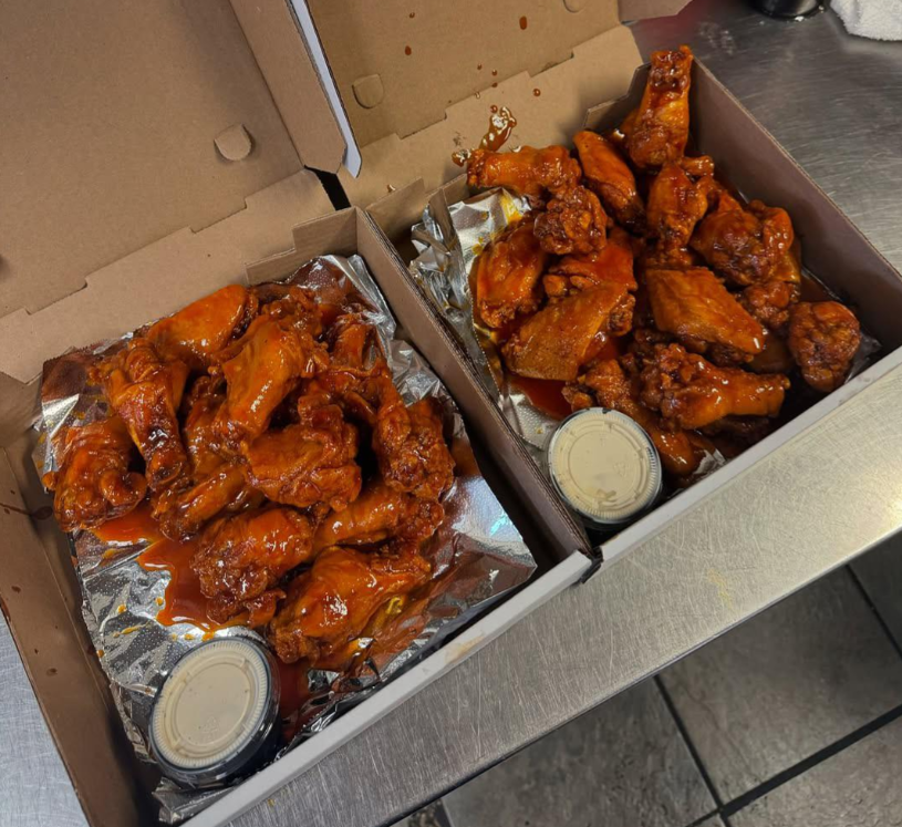 Two open pizza boxes on a metal counter, each filled with saucy buffalo chicken wings and a selection of wing sauces on foil, with a small container of dipping sauce in each box.