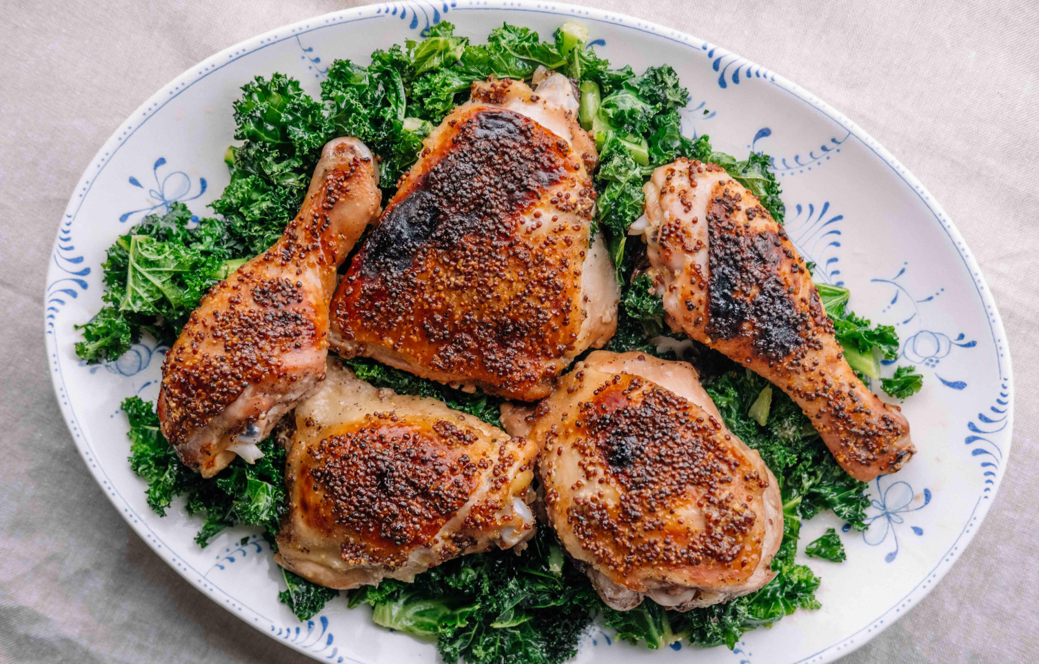 Roasted chicken thighs and drumsticks with a mustard seed crust are served on a bed of cooked kale, inspired by classic wing sauces, on a white and blue patterned oval platter.