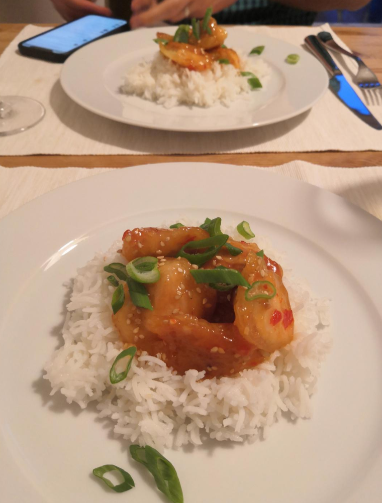 Two plates of white rice topped with saucy shrimp—coated in flavorful wing sauces—and sliced green onions sit on a table. A person in the background is holding a phone and eating, with forks, knives, and a wine glass also visible.