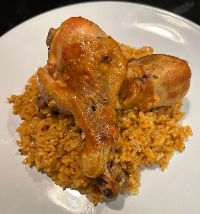 Two cooked chicken drumsticks served on a bed of seasoned, orange-colored rice on a white plate, with flavors reminiscent of popular wing sauces. The dish appears to be flavorful and well-cooked.