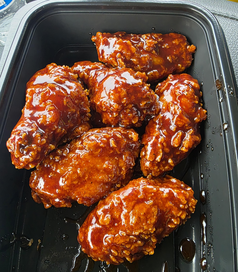 Six crispy fried chicken wings coated in a glossy, dark reddish-brown barbecue wing sauce are arranged in a black plastic takeout container.