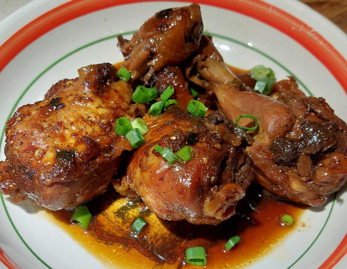 Braised chicken drumsticks glazed in a dark, glossy wing sauce, garnished with chopped green onions, served on a white plate with a red and green border.