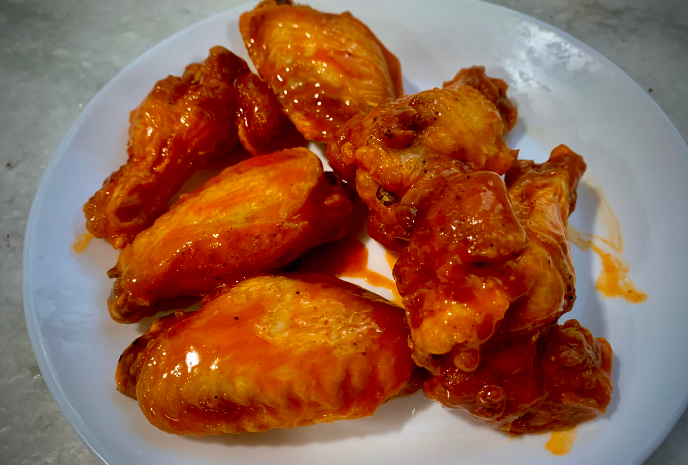 A white plate holds six saucy buffalo chicken wings, each coated in a shiny, orange-red glaze—one of the most popular wing sauces—sitting invitingly on a light-colored surface.