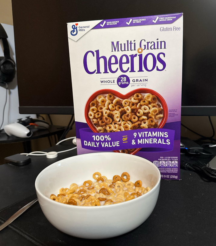 A bowl of Multi Grain Cheerios cereal with milk sits in front of a Cheerios cereal box on a desk, surrounded by computer monitors and headphones—showcasing how everyday products fit seamlessly into modern routines.