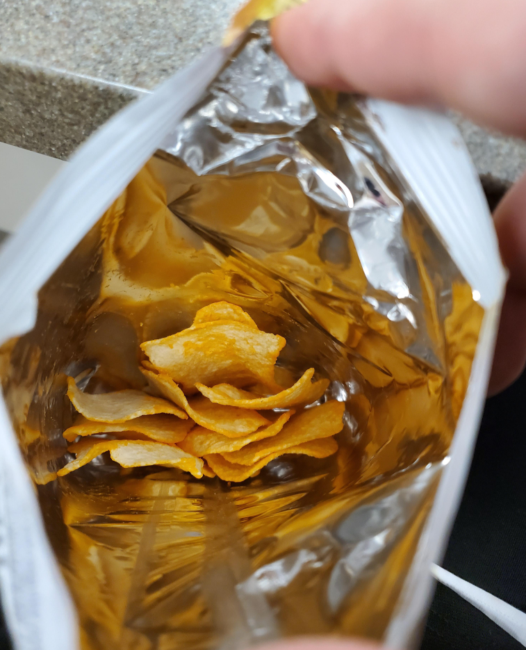 A hand holds open a bag of potato chips, revealing only a small amount of chips at the bottom and a lot of empty space inside the shiny, foil-lined bag—a familiar sight with many everyday products.