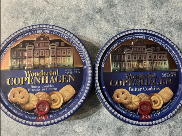 Two blue tins of Wonderful Copenhagen butter cookies are shown on a marble surface. The tins feature images of butter cookies and a colorful Copenhagen building facade. The left tin is 454g, and the right tin is 340g.