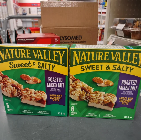 Two Nature Valley Sweet & Salty Roasted Mixed Nut granola bar boxes sit on a store counter, one with 5 bars (175g) and one with 6 bars (210g). Shelves and a brown box are visible in the background.