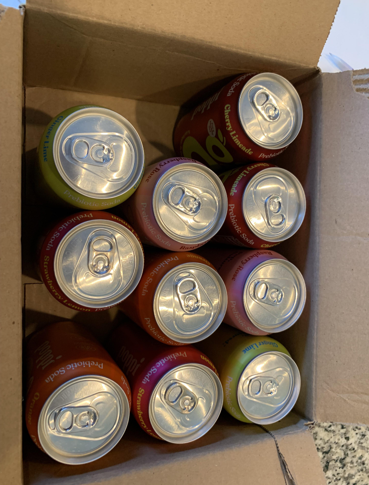 Nine cans of sparkling water with multicolored labels, a staple among everyday products, are arranged in a cardboard box and viewed from above. The cans have pull tabs, and the box rests on a speckled countertop.